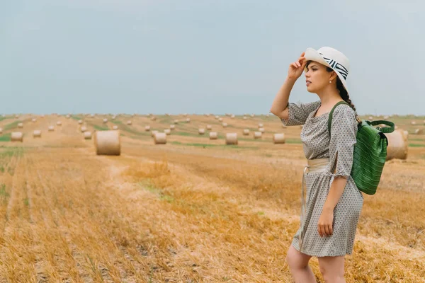 Menina bonita em um chapéu e com uma mochila no meio de um campo de trigo com fardos em uma noite de verão — Fotografia de Stock