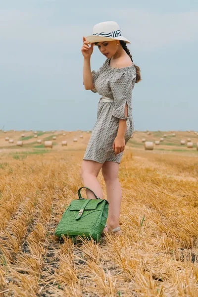 Bella giovane donna in un cappello e con uno zaino in mezzo a un campo di grano con balle in una sera d'estate — Foto Stock
