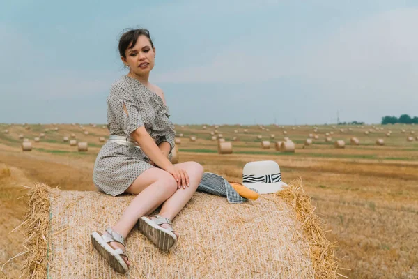 Menina bonita sentada no topo do fardo no meio do campo de trigo com fardos na noite de verão — Fotografia de Stock