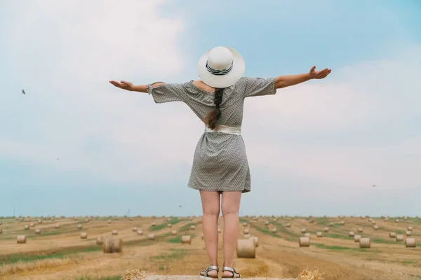 Mulher bonita em um chapéu olha para o céu com as mãos até o céu no meio de um campo de trigo com fardos na noite de verão — Fotografia de Stock
