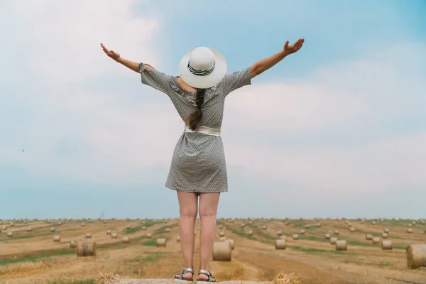 Jovem mulher em um chapéu olha para o céu com as mãos até o céu no meio de um campo de trigo com fardos na noite de verão — Fotografia de Stock