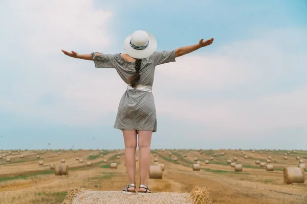 Mulher bonita em um chapéu olha para o céu com as mãos até o céu no meio de um campo de trigo com fardos na noite de verão — Fotografia de Stock