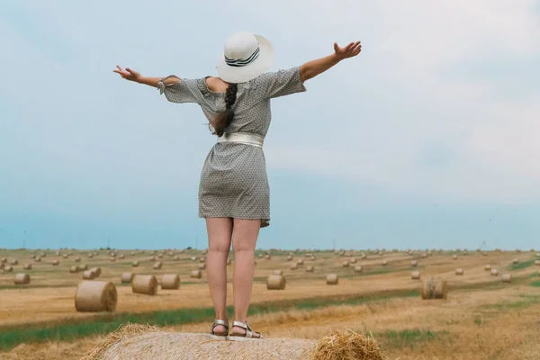 Jovem mulher em um chapéu olha para o céu com as mãos até o céu no meio de um campo de trigo com fardos na noite de verão — Fotografia de Stock