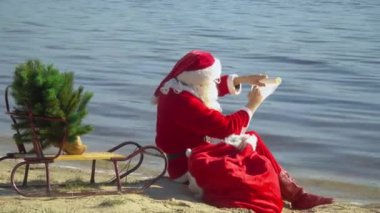 Noel Baba elinde hediye çantasıyla gölün kumlu kıyısına oturur ve listeye bir şeyler yazar.