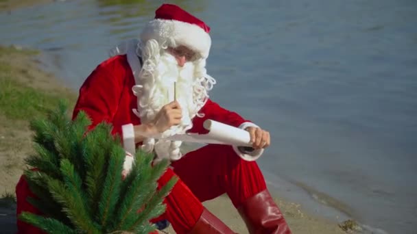 산타클로스는 호수의 모래사장에 선물 가방을 들고 앉아서 아래에 무엇인가를 적는다 — 비디오