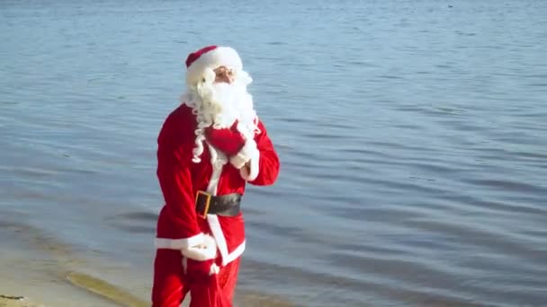 Babbo Natale si trova vicino al mare o al lago con una borsa di regali — Video Stock