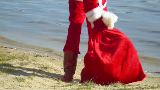 Noel Baba bir torba hediyeyle okyanusun kenarında duruyor. Noel Baba, içinde hediyeler olan bir çantayı kuma koyar. Bacakları gibi.. — Stok video
