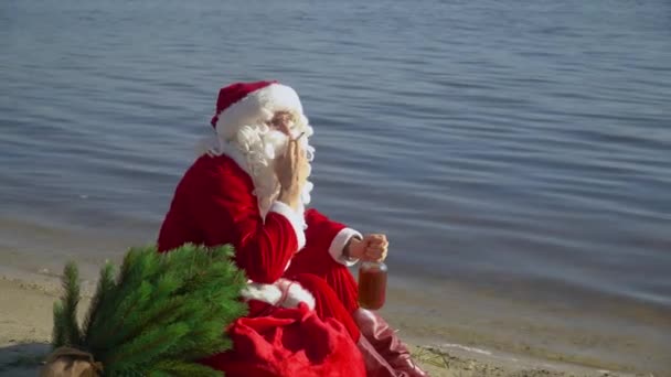 Bad Santa Claus zit met een kadozakje aan de zandige oever van een meer en rookt een sigaret terwijl hij een fles alcohol vasthoudt. Dronken Kerstman. — Stockvideo