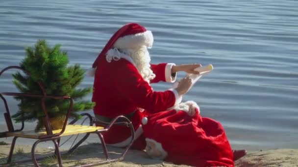 Santa Claus se sienta con una bolsa de regalo en la orilla arenosa del lago y escribe algo en la lista — Vídeo de stock