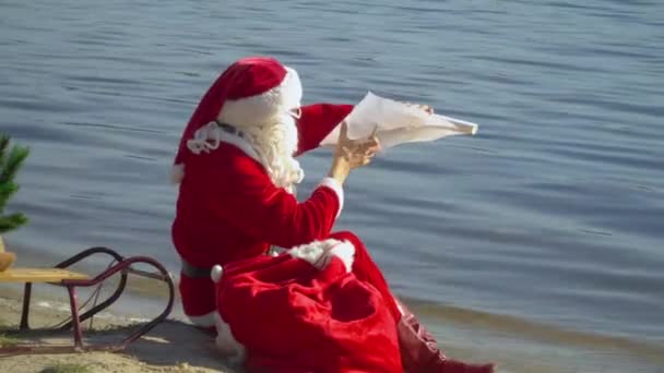 Papai Noel senta-se com um saco de presente na costa arenosa do lago e lê a lista — Vídeo de Stock