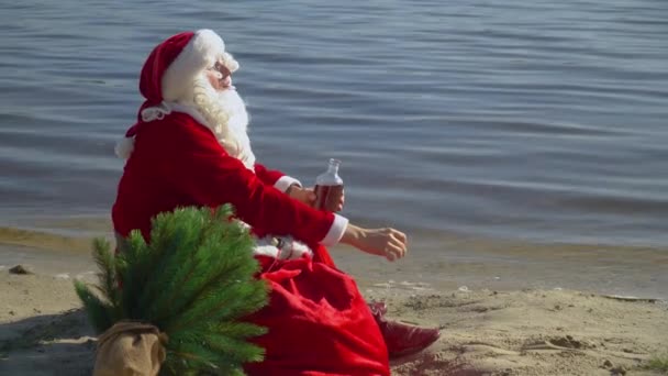Jultomten sitter med en påse presenter på sandstranden av ett hav och röker en cigarett och dricker alkohol från en flaska. Fylld jultomte — Stockvideo