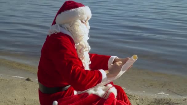 Papai Noel senta-se com um saco de presentes na costa arenosa do lago e depois de hesitação escreve algo para a lista — Vídeo de Stock