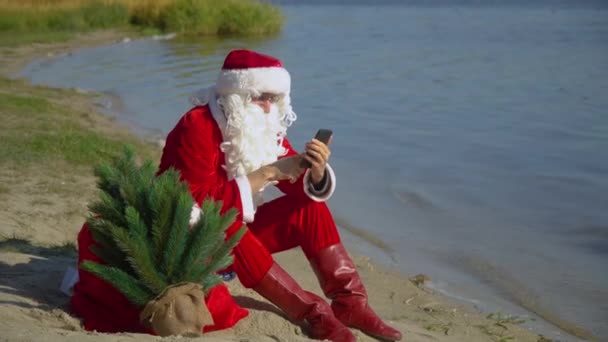 Mikulás ül egy zsák ajándékot a homokos parton a tó és működik az okostelefon — Stock videók