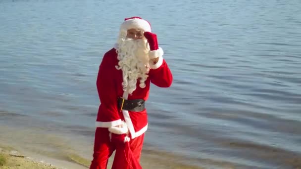Santa Claus se encuentra junto al río o lago con una bolsa de regalos — Vídeos de Stock