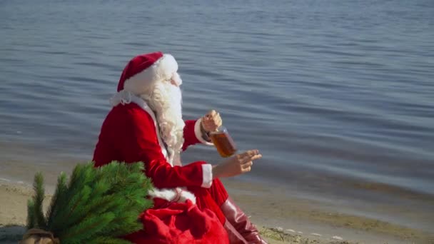 Der böse Weihnachtsmann sitzt mit einem Sack voller Geschenke am sandigen Ufer eines Sees, raucht eine Zigarette und trinkt Alkohol aus einer Flasche. Betrunkener Weihnachtsmann — Stockvideo