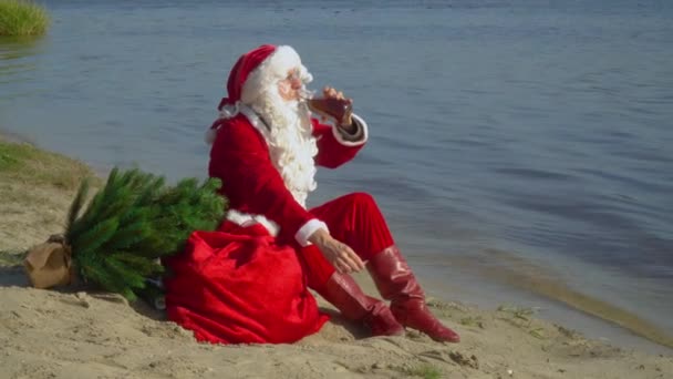 Papai Noel senta-se com um saco de presentes na costa arenosa de um lago e fuma um cigarro e bebe álcool de uma garrafa. Papai Noel bêbado — Vídeo de Stock