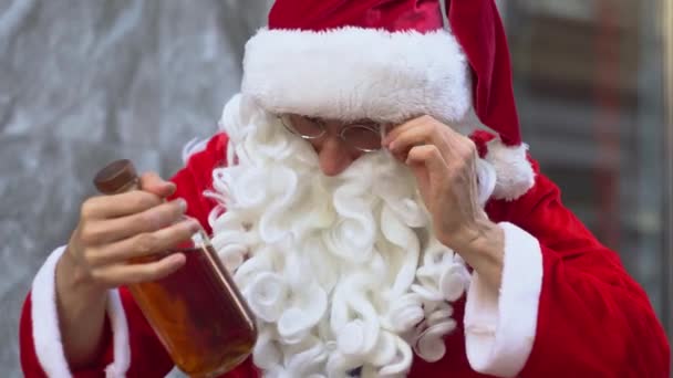 Поганий п'яний Санта Клаус сидить біля будівлі міського офісу і знаходить пляшку алкоголю. Санта Клаус читає етикетку алкоголю і показує жест "ОК" рукою. — стокове відео