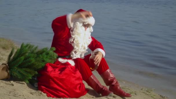 Santa Claus sitzt am sandigen Ufer des Sees und riecht den Geruch von Alkohol und Flaschen. Betrunkener Weihnachtsmann — Stockvideo