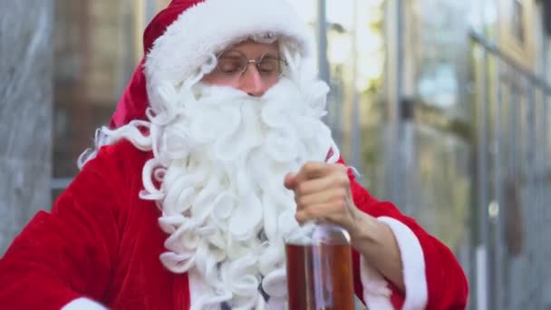 Babbo Natale ubriaco cattivo si siede vicino a un edificio per uffici della città. Babbo Natale beve alcol da una bottiglia per strada — Video Stock