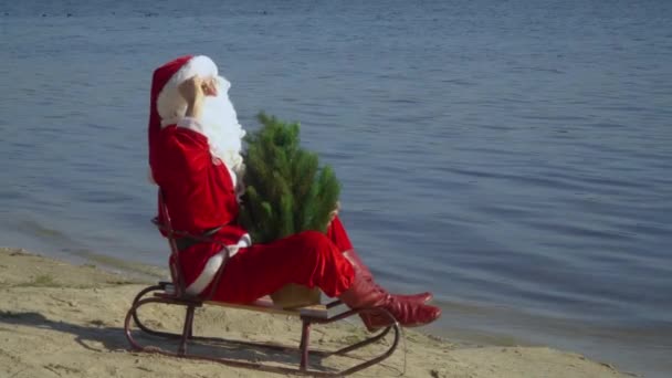 Santa Claus se sienta en un trineo en la orilla arenosa de un lago, sostiene un árbol de Navidad y mira a la distancia. Santa en las Maldivas — Vídeos de Stock