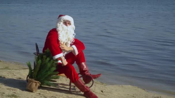 Papai Noel senta-se em um trenó na costa arenosa do lago, corrige sua barba, óculos e boné olhando para a câmera — Vídeo de Stock