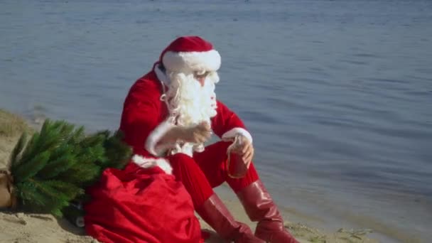 Santa Claus se sienta en la orilla arenosa del lago oliendo el olor del alcohol y sonríe a la cámara. Borracho Santa Claus — Vídeos de Stock