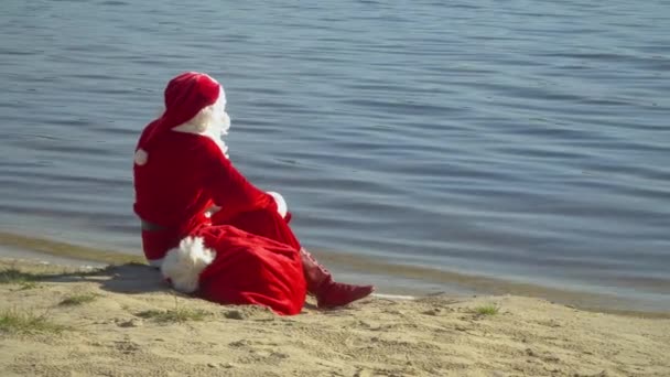 Papai Noel senta-se junto ao rio ou lago com um saco de presentes. Papai Noel olha para a distância na água. Santa relaxante — Vídeo de Stock