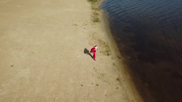 Santa Claus se encuentra con una bolsa de regalo en la orilla arenosa del lago. Santa Claus corrige su sombrero. Santa en los trópicos. Vista superior — Vídeos de Stock