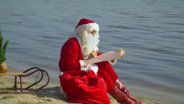 De Kerstman zit met een zak geschenken op de zandkust van het meer en reflecteert op zijn cadeaulijst. Kerstman in het resort — Stockvideo