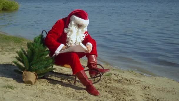 Papai Noel senta-se em um trenó na costa arenosa de um lago e escreve algo no papel. Santa no mar — Vídeo de Stock