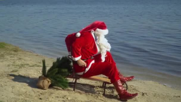 Der Weihnachtsmann sitzt im Schlitten am sandigen Ufer des Sees und versucht, auf dem Schlitten näher ans Wasser zu kommen. Weihnachtsmann auf dem Meer — Stockvideo