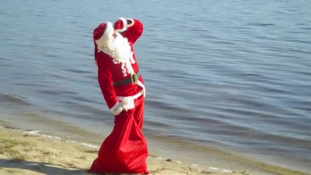 De kerstman staat bij de rivier of het meer met een zak cadeautjes. De Kerstman kijkt naar de lucht — Stockvideo