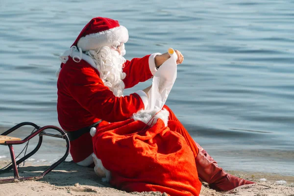 Noel Baba bir hediye çantasıyla gölün kumlu sahilinde oturur ve bir liste okur. — Stok fotoğraf