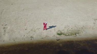Noel Baba, elinde hediye çantasıyla gölün kumlu kıyısında duruyor. Noel Baba kameraya merhaba ve Ok işaretlerini gösterir. Üst Manzara. Noel Baba tropiklerde