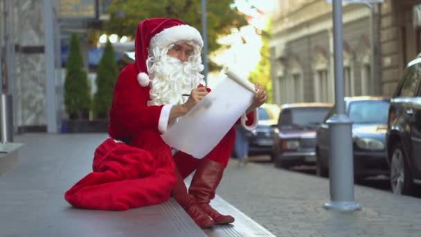 Babbo Natale si siede vicino all'edificio degli uffici della città e scrive qualcosa su un pezzo di carta — Video Stock