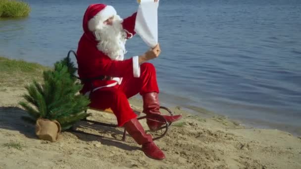 Papai Noel senta-se em um trenó na costa arenosa de um lago e lê algo de um pedaço de papel — Vídeo de Stock