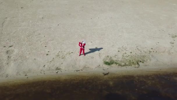 De Kerstman staat met een kadozakje aan de zanderige oever van het meer. De Kerstman toont hallo en OK gebaren naar de camera. Bovenaanzicht. Kerstman in de tropen — Stockvideo