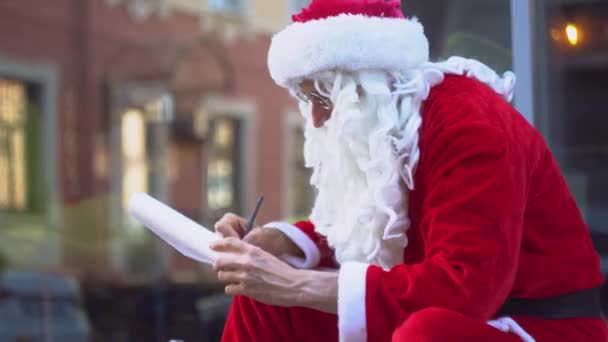 Санта Клаус сидить біля будівлі міського офісу і щось пише на клаптику паперу. — стокове відео