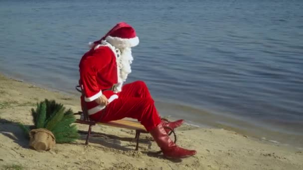 Санта Клаус сидит в санях на песчаном берегу озера и пытается продвинуться на санях ближе к воде. Санта на море — стоковое видео