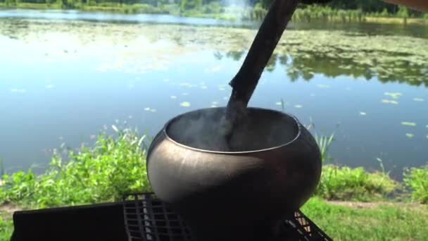 Залізний литий котел кипить вухо над відкритим колодним вогнем на фоні озера — стокове відео