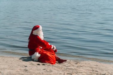 Noel Baba elinde hediye çantasıyla gölün kumlu kıyısına oturur ve gölete bakar.
