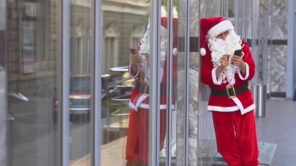 Weihnachtsmann steht an der Wand eines Bürogebäudes und bedient sich eines Smartphones — Stockvideo