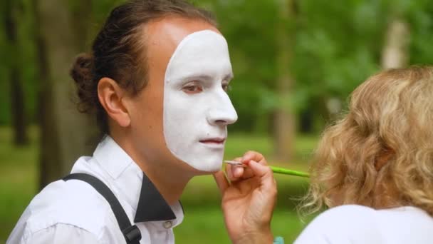 Девушка рисует лицо молодому человеку пантомиму белым карандашом — стоковое видео