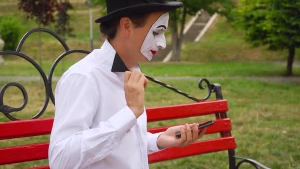 Grappige mime zit op een bankje in het park met een telefoon — Stockvideo