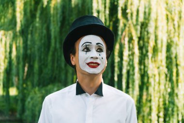 Parkta yeşil arka planda gülümseyen siyah şapkalı pandomimci.