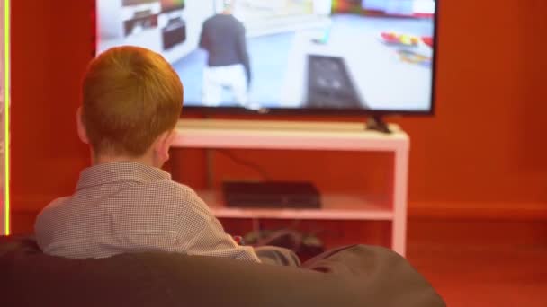 Un chico pelirrojo se sienta frente a una gran pantalla y sostiene un joystick en sus manos. El niño juega un juego de realidad virtual — Vídeos de Stock