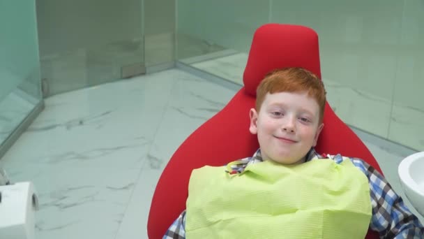 Счастливый рыжеволосый мальчик на приеме у дантиста в стоматологическом кресле, который встает. Безопасная стоматология — стоковое видео