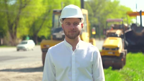 Portret van de manager van de reparatiewerkzaamheden in een witte helm tegen de achtergrond van een wegenbouwplaats — Stockvideo