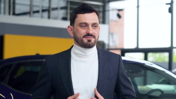 Elegante hombre está de pie cerca del coche en la sala de exposición de coches. Auto negocio, venta de coches, tecnología y concepto de personas — Vídeos de Stock