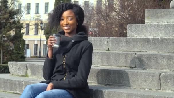 Feliz jovem afro-americana bebendo café de xícara de papel sentado na escada de rua. Materiais ambientais. Bebidas e conceito de pessoas. Fim da quarentena — Vídeo de Stock
